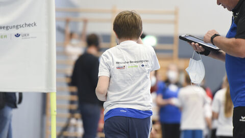 Hessischer Bewegungscheck in der Umsetzung in einer Sporthalle: von hinten ist ein Schuljunge in Sportkleidung zu sehen, rechts von ihm ein Betreuer vom Sportkreis der den Bewegungscheck mit den Kindern durchführt.