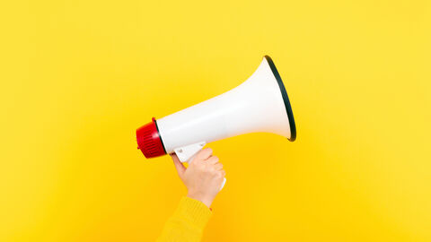 Vor einem gelben Hintergrund ist ein rot-weißes Megaphon zu sehen, das von einer Hand mit gelben Ärmel hochgehalten wird.