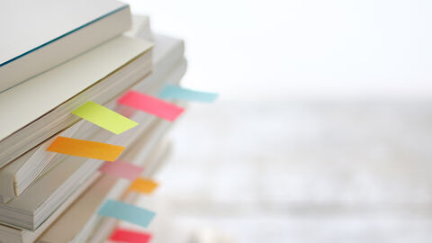 Qualitätssinstrumente Vor einem weißen bzw. hellgrauen Hintergrund sieht man links im Bild eine Nahaufnahme eines Stapels verschiedener weißer Hefte bzw. Bücher, bei denen farbliche Seitenmarker herausschauen.