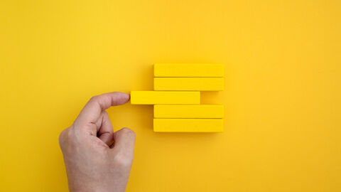 Vor einem gelben Hintergrund liegen 5 gelbe Holzbausteine übereinander. Der mittlere Baustein ist etwas nach links verlagert. Links von den Bausteinen ist eine Hand zu sehen, die mit dem Zeigefinger den herausschauenden Baustein wieder gerade zurückschieben möchte.