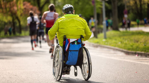 Auf einer autofreien Straße ist von hinten ein Mann mit hellgrüner Sportjacke und Helm in einem Rollstuhl zu sehen. Im Hintergrund sind weitere Läufer von hinten zu erkennen.