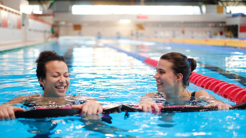 Zwei junge Frauen halten sich im Schwimmbad an Schwimmnudeln fest und lächeln sich dabei gegenseitig an. Im Hintergrund ist ein Hallenbad zu erkennen.