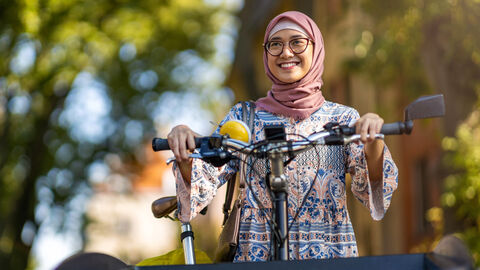 Eine Frau mit Kopftuch und Brille hält sich mit beiden Händen lächelnd an einem Fahrrad fest. Im Hintergrund sind verschwommen Bäume und ein blauer Himmel zu erkennen. 