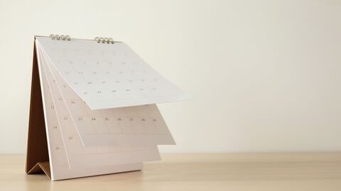 Auf einem Holztisch und vor beigem Hintergrund steht ein umklappbarer Tischkalender mit einer Monatsansicht.