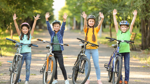 Vier junge Kinder, davon zwei Mädchen und zwei Jungs, stehen jeweils neben ihren Fahrrädern, die nebeneinander in einer Reihe aufgestellt sind. Dabei halten sie alle die Hände hoch und lächeln in die Kamera. Alle vier Kinder tragen Fahrradhelme. Im Hintergrund ist ein Kiesweg bzw. -allee zu sehen und rechts und links davon Bäume.
