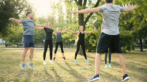In einem Park sind sechs springende junge Erwachsene in Sportkleidung zu sehen. Beine und Arme sind hierbei weit auseinandergestreckt. Vorne rechts ist von hinten der vermutliche Übungsleiter zu sehen, alle weiteren Erwachsenen schauen ihn hierbei lächelnd an. 