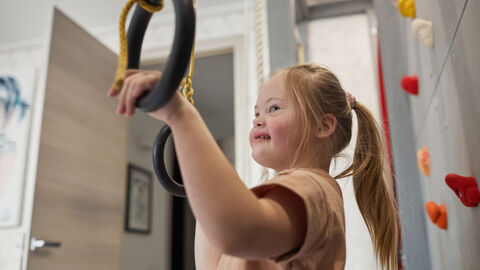 Ein junges Mädchen, vermutlich mit Trisomie 21, ist vom Profil in einer Sporthalle zu sehen. Sie hält sich mit beiden Händen an Turnringen fest und lächelt. Rechts von ihr ist eine Kletterwand zu sehen.