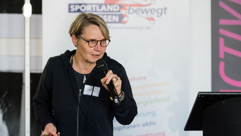 Impulsvortrag von Dr. Katrin Korth, Korth StadtRaumStrategien Dr. Katrin Korth während Ihres Impulsvortrages mit Mikro in der Hand, im Hintergrund sind Roll-Ups zu sehen