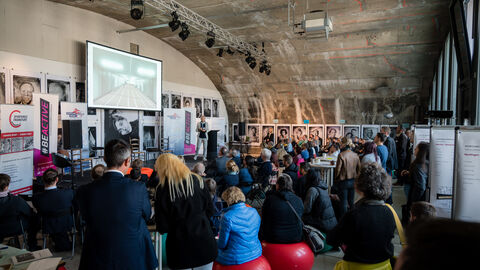 Bewegte Bestuhlung  Hintenansicht vom voll besetzten Veranstaltungsraum, teilweise sitzen die Teilnehmenden auf Sitzbällen. Im Hintergrund ist die Bühne mit der Leinwand und Dipl.-Ing. Dirk Schelhorn während seines Vortrages zu sehen.