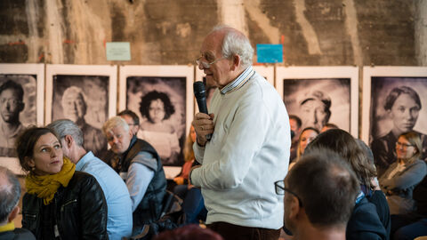 Fragerunde mit den Teilnehmenden Nahaufnahme auf das Publikum während der offenen Fragerunde, hier ist von der Seitenansicht ein Teilnehmer zu sehen der mit Mikro in der Hand aufgestanden ist und links Richtung Bühne blickt.