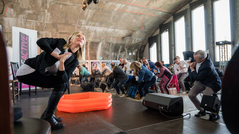Bewegte Pause Seitenansicht links von der Übungsleiterin und rechts vom Publikum während einer Bewegungsübung, bei der sich alle in der Hockenposition nach rechts drehen und hierbei die Hände vor der Brust halten.