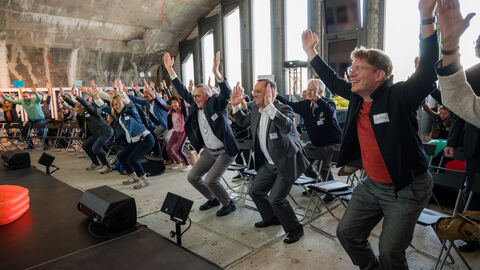 Bewegte Pause Seitenansicht von lachenden Teilnehmenden während einer Bewegungsübung, bei der alle in der Hocke die Arme nach oben strecken.