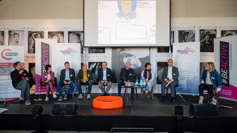 Intersektoral besetzte Diskussionsrunde rund um Anliegen der Bevölkerung und der Teilnehmenden Intersektoral besetzte Diskussionsrunde rund um Anliegen der Bevölkerung und der Teilnehmenden. Hier sind alle acht Podiumsgäste sitzend auf der Bühne zu sehen, links von ihnen sitzt die Moderatorin. In der Mittle liegt ein Planschbecken, im Hintergrund ist die Leinwand sowie eine Fotoausstellung zu erkennen.