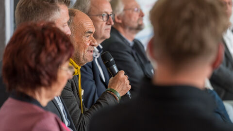 Intersektoral besetzte Diskussionsrunde rund um Anliegen der Bevölkerung und der Teilnehmenden Intersektoral besetzte Diskussionsrunde rund um Anliegen der Bevölkerung und der Teilnehmenden. Hier von der Seitenansicht Roland Frischkorn scharf gestellt zu sehen, er schaut rechts in Richtung des Publikums. Rechts und links von ihm sitzen weitere Podiumsgäste.