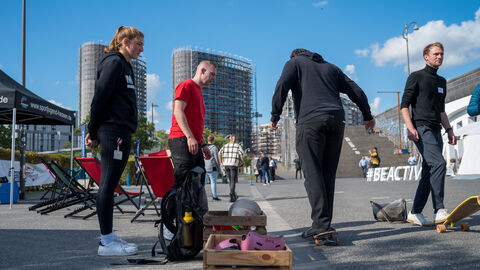 Vielfältiges Bewegungsprogramm auf dem Hafenparkgelände zum Mitmachen: Sportmobil Adventure Cube (Sportjugend Hessen) Vielfältiges Bewegungsprogramm auf dem Hafenparkgelände zum Mitmachen: Sportmobil Adventure Cube (Sportjugend Hessen). Hier probiert zwei Teilnehmer das Skateboarden aus, umgeben von Personal der Sportjugend Hessen.