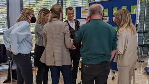 Teilnehmende bei der Bilderrätsel-Methode zum Kennenlernen der Good Practice-Kriterien. Hier sieht man Teilnehmende von hinten vor einer Pinnwand stehen an der das Bilderrätsel hängt.