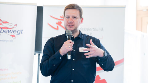 Fachforum 2: bewegt bleiben in der Kommune, Praxisbeispiel Stefan Burger Stefan Burger während der Vorstellung des Praxisbeispiels mit Mikro in der Hand, im Hintergrund sind Roll-Ups zu sehen.