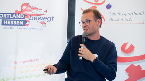 Fachforum 2: bewegt bleiben in der Kommune, Praxisbeispiel Jörg Peters Jörg Peters während der Vorstellung des Praxisbeispiels mit Mikro und Laserpointer in der Hand, im Hintergrund sind Roll-Ups zu sehen.