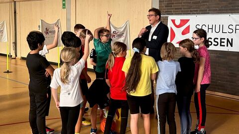 Staatssekretär Stefan Sauer steht in einer Sporthalle. Um ihn herum stehen viele Kinder, mit denen er redet. Einige Kinder melden sich. Auf dem Boden sind die klassischen Markierung einer Sporthalle. Im Hintergrund ist ein Banner zu sehen. 