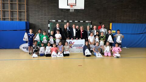 Gruppenbild der Auftaktveranstaltung. Alle Personen sind in Reihen abgebildet. Zu sehen sind unter anderem eine dritte Klasse mit vielen Jungen und Mädchen, sowie Vertretungen aller Projektpartner. Staatssekretär Stefan Sauer hält ein Banner mit dem Logo des Bewegungschecks. Im Hintergrund sind Turnmatten und ein Basketballkorb zu sehen.