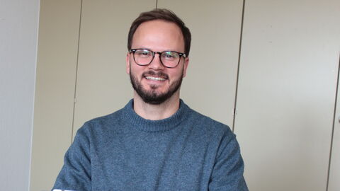 Portrait des Bewegungskoordinators im Vogelsbergkreis Dominic Günther. Er lächelt in die Kamera und sitzt vor seinem aufgeklappten Tablet. Hinter ihm ist eine Büroschrankwand zu sehen.