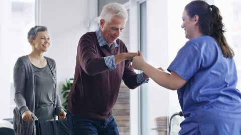 Pflegerin hilft älterem Herr aus dem Rollstuhl. Sie steht vor ihm und hält beide Hände Der Herr mit grauen Haaren steht bereits fast vollständig und lächelt. Der Rollstuhl wird von einer dritten weiblichen Person festgehalten.  Auch sie lacht. 