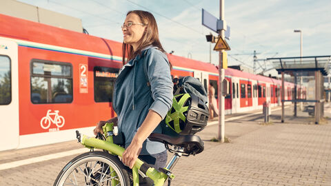 Bei schönem Wetter ist eine Frau mit eingeklappten Fahrrad auf einem Bahngleis zu sehen. Im Hintergrund steht ein eingefahrener roter Regionalzug.