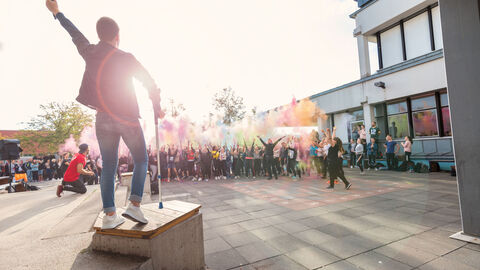 Mann steht auf Podest mit Mikrofon in der Hand und hebt die Hand in die Luft. Vor ihm stehen viele Menschen und werfen buntes Pulver in die Luft