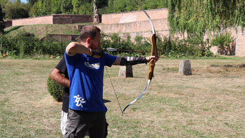 Mann spannt Pfeil und bogen. Trainer steht hinter ihm.