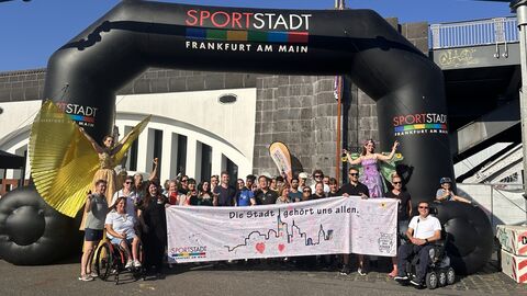 Gruppe von Menschen, die ein Banner hochhalten unter einem aufblasbaren Torbogen des Sportamts. 