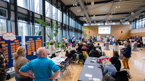 Aufmerksame Zuhörerinnen und Zuhörer: Teilnehmende lauschen den Moderatorinnen bei der Erläuterung zum Ablauf Bild der Teilnehmenden vom Ende des Raums. Meschen hören den Moderatorinnen zu.