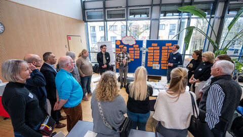 "Rundgang durch die bewegungsfreundliche Kommune" in Gruppen: Teilnehmende lauschen der Vorstellung der Lebensweltexperten und -expertinnen bei der Vorstellung der Lebenswelt Schule Menschen stehen vor Pinnwand und hören Redner zu