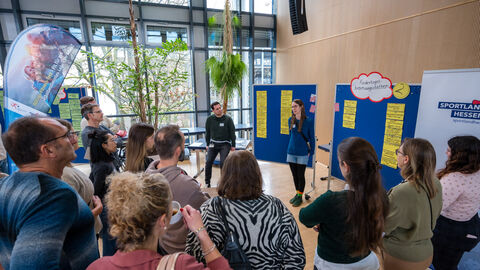 "Rundgang durch die bewegungsfreundliche Kommune" in Gruppen: Teilnehmende lauschen der Vorstellung der Lebensweltexperten und -expertinnen bei der Vorstellung der Lebenswelt Kindertagesbetreuungsstätte  Menschen stehen vor Pinnwand und hören Rednerin zu