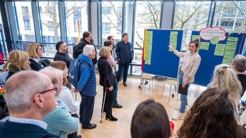 "Rundgang durch die bewegungsfreundliche Kommune" in Gruppen: Teilnehmende lauschen der Vorstellung der Lebensweltexperten und -expertinnen bei der Vorstellung der Lebenswelt Pflege- und Senioreneinrichtungen  Menschen stehen vor Pinnwand und hören Rednerin zu
