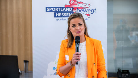 Moderatorin Béatrice Frank (HAGE) kündigt den nächsten Programmpunkt an Frau mit Mikrofon in der Hand vor Roll-up des Landesprogramms