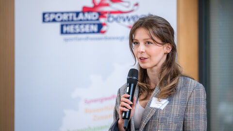 Moderatorin Lena Ondrasch (HAGE) bei der Ankündigung des nächsten Programmpunktes Frau mit Handmikrofon vor Roll-up des Landesprogramms