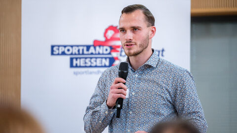Marius Hamel (Bewegungskoordinator im Landkreis Waldeck-Frankenberg) bei der Vorstellung eines Beispiels guter Praxi Mann mit Mikrofon in der Hand vor Roll-up