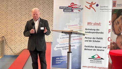 Mann mit Handmikrofon neben Stehpult vor verschieden Bannern 