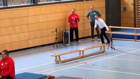 Mann nimmt Anlauf für die Rennstrecke in der Sporthalle. Weitere Zuschauer stehen drum herum 