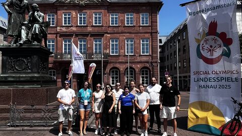 Eine Gruppe von elf Personen steht vor einem historischen roten Backsteingebäude mit einem Uhrenturm bei strahlend blauem Himmel. Links im Bild ist eine Bronzestatue von zwei Personen auf einem Sockel zu sehen. Rechts im Bild befindet sich ein großes Banner mit dem Text ‚#gemeinsamstark SPECIAL OLYMPICS LANDESSPIELE HANAU 2025‘. Die Personen wirken fröhlich und sind lässig gekleidet, einige tragen Event-Ausweise oder Sportkleidung.